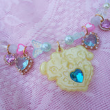 A close-up of a pastel beaded charm bracelet or necklace laid on pink textured fabric. The central charm is a pale yellow bear-shaped pendant with a bright blue heart gem, surrounded by translucent pearls, pastel beads, butterfly-shaped beads, and heart-shaped charms with blue and pink stones in gold-toned settings.