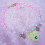 A pastel pink beaded necklace with a yellow cartoon bear pendant, decorated with iridescent heart beads, pink and blue crystals, and gold-tone hardware, displayed on a pink lace fabric background.